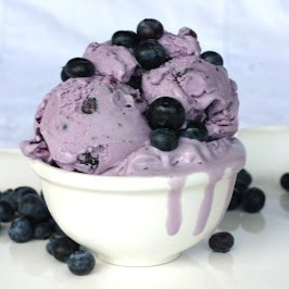 A bowl of blueberry ice cream in a white bowl, with melty purple ice cream drips running down the side. The ice cream is topped with fresh blueberries and there are more blueberries scattered around the table surface below.