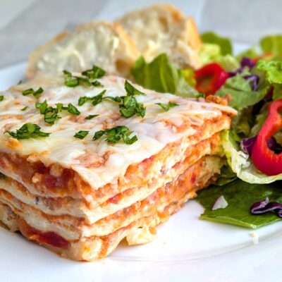 A serving of lasagna on a white plate with a side salad and buttered bread slices.