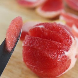 A segment of grapefruit on a knife, with the whole peeled grapefruit on a cutting board in the background.
