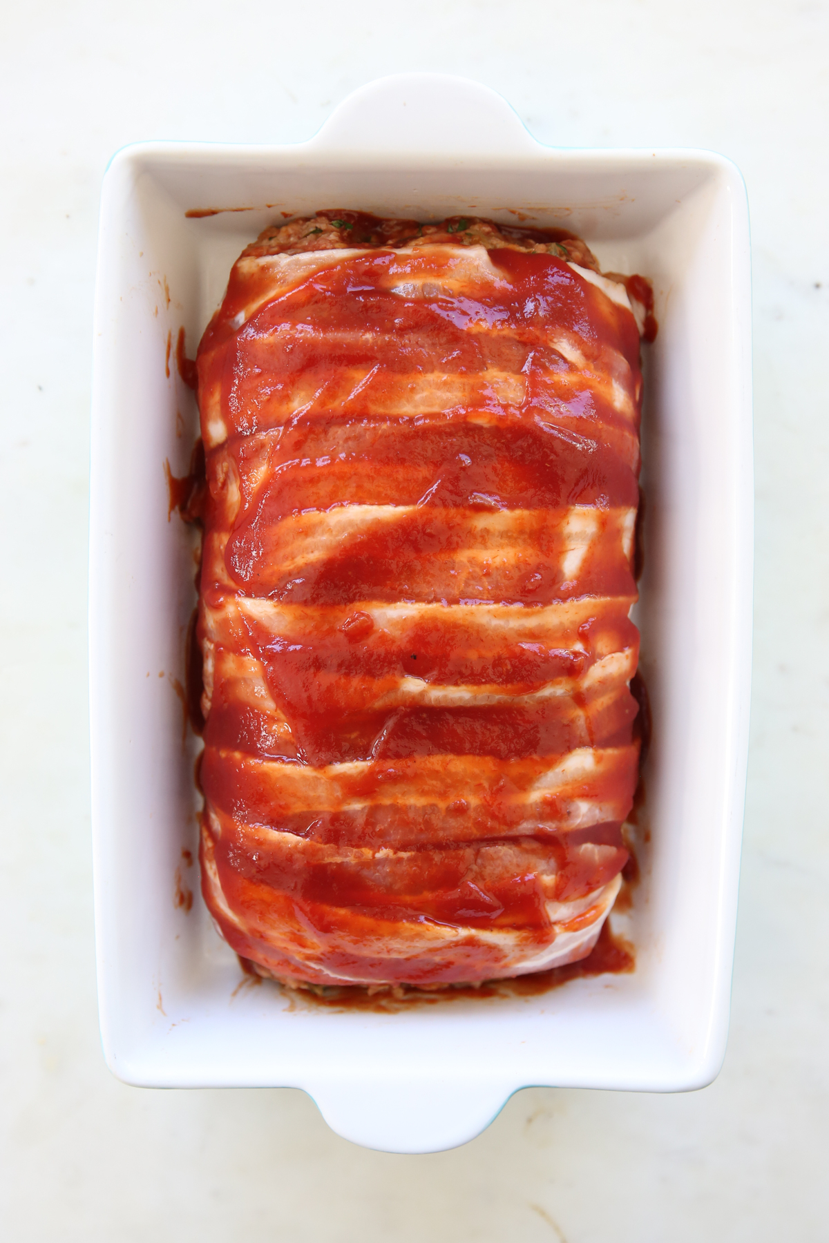 meatloaf wrapped in bacon spread with sauce, in a baking pan