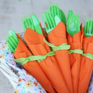 A basket filled with cute utensil bundles that look like carrots. Green utensils are wrapped in orange napkins with a little green bow on each.