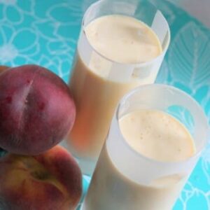 Two glasses filled with Peach Milkshakes, next to a pile of fresh peaches.