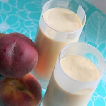 Two glasses filled with Peach Milkshakes, next to a pile of fresh peaches.