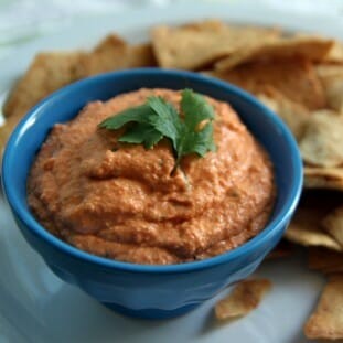 A small bowl filled with toasted red pepper hummus, surrounded by pita chips.