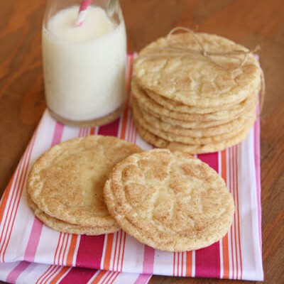 A couple of loose snickerdoodles sit on a pink, red, and white striped cloth, with a stack of snickerdoodles and a glass of milk in the background.