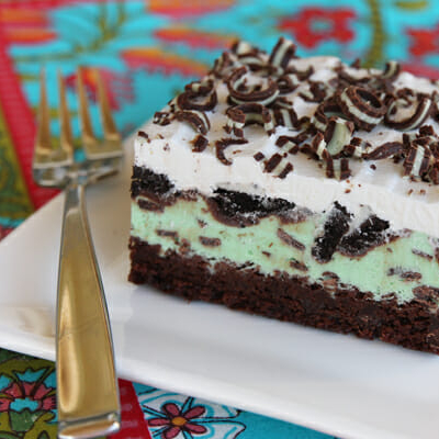 A square of mint chocolate chip ice cream brownie dessert with layers of brownie, ice cream, ganache, whipped topping, and Andes mints, on a white plate with a fork.