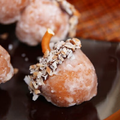 An acorn treat made from a doughnut hole, with the top dipped in chocolate and then toasted coconut, with a curved pretzel piece as a stem.