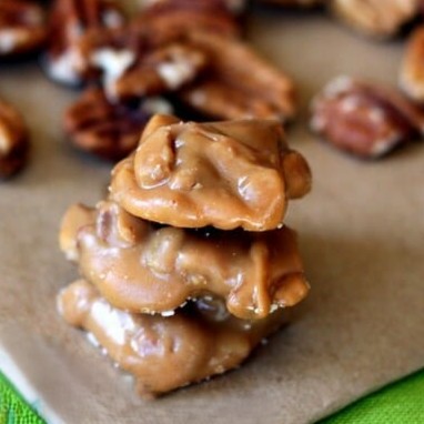 A stack of 3 little pralines on a baking sheet, with pecans scattered in the background.
