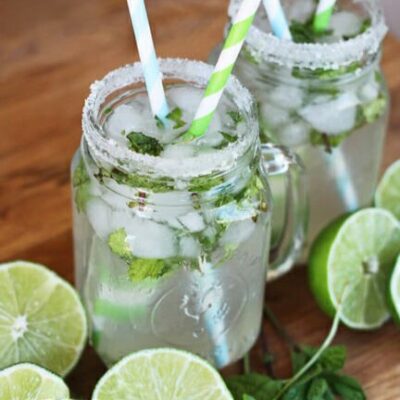 Two mason jar mugs full of iced mint limeade, surrounded by halved limes and loose mint.