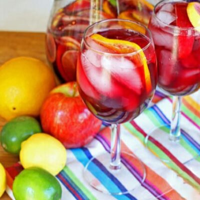 A red sangria with no alcohol in two wine glasses, surrounded by colorful fruits on a colorful striped napkin.