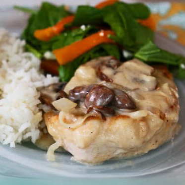 A pork chop, smothered in brie, inions, and mushrooms, sits on a plate with a side of rice and a salad.