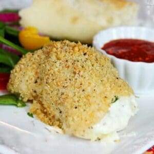 A chicken breast, stuffed with spinach and cheese, coated in seasoned bread crumbs sits on a white plate with a side of tomato sauce, vegetables, and bread.