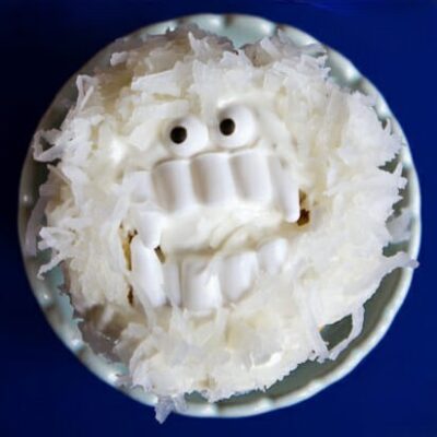 A yeti cupcake, made by adding vanilla frosting, shredded coconut, candy eyes, and plastic vampire teeth.