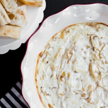 A pie plate full of hot French onion dip. Pieces of bread are on another plate in the background.