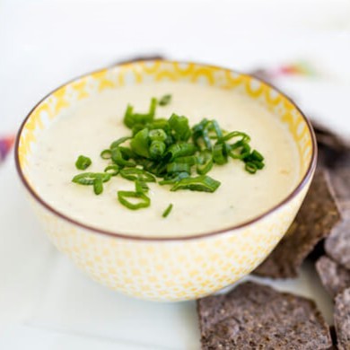 A bowl of queso, topped with green onions, next to a bed of blue corn tortilla chips.