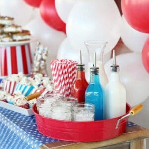A patriotic Italian Soda Bar spread with flavored syrups, glasses full of ice, cute straws, balloons, and treats.