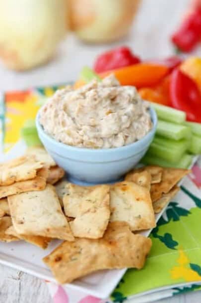 A bowl of caramelized onion dip, surrounded by fresh cut up veggies and pita chips.