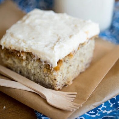 A square slice of banana cake, with a cream cheese frosting, sitting on a piece of brown parchment next to a couple of disposable bamboo forks. 