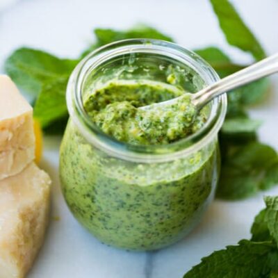 A little glass pot of mint-basil pesto, surrounded by basil leaves and hunks of parmesan cheese.
