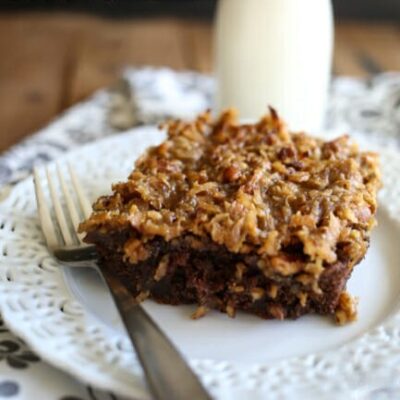 A slice of coconut crunch topped chocolate cake on a white plate with a bottle of milk in the background.