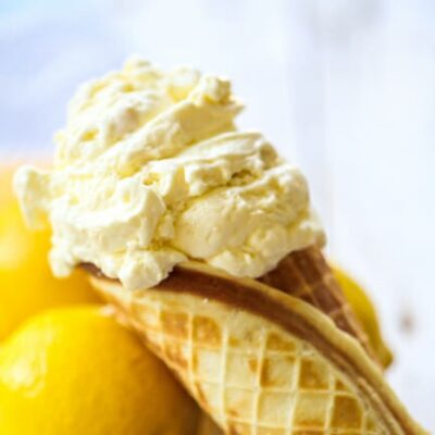 A scoop of light yellow lemon custard in a homemade waffle cone, leaning on whole, fresh lemons.