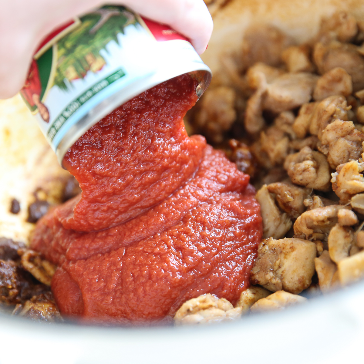 a hand pouring tomato sauce out of a can into a pot of chicken