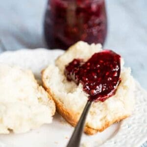 A fluffy biscuit on a white plate. The biscuit has been pulled in half and has a small spoon filled with fresh raspberry jam laid on top of it.