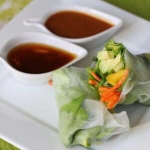 A spring roll, wrapped in butter lettuce and rice paper, cut in half to reveal fresh veggie filling. Two dipping sauces accompany the fresh spring roll.