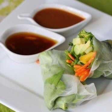 A spring roll, wrapped in butter lettuce and rice paper, cut in half to reveal fresh veggie filling. Two dipping sauces accompany the fresh spring roll.